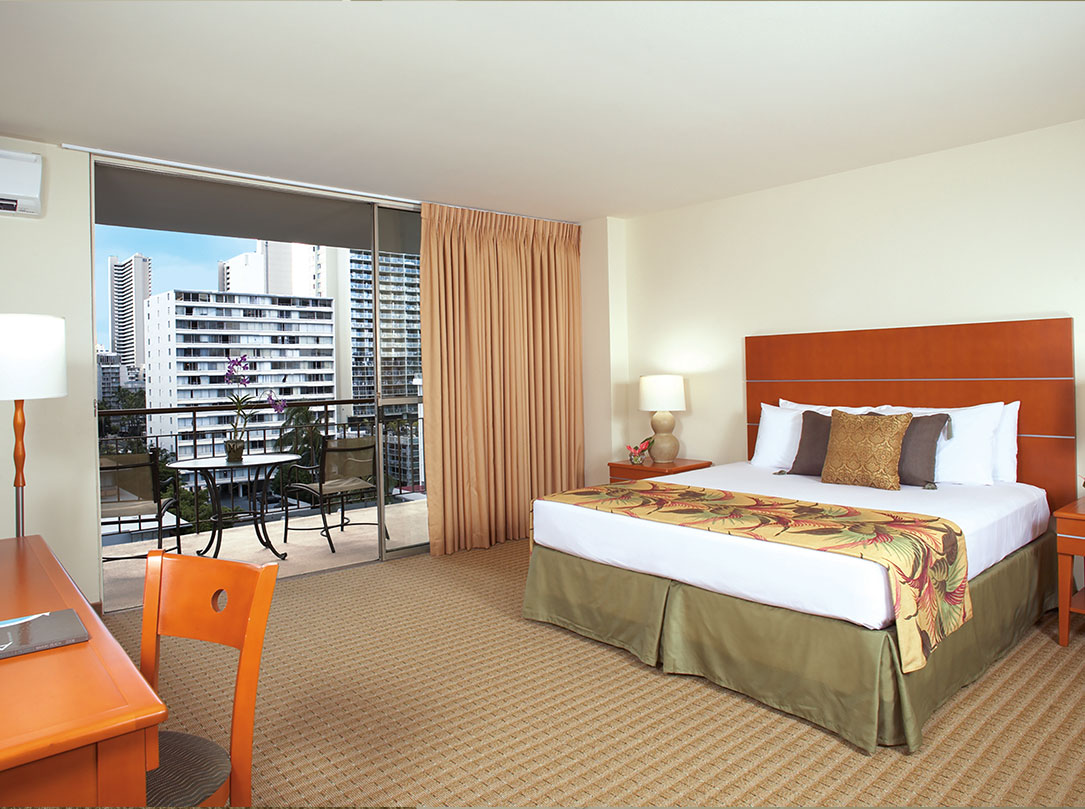Bedroom with Queen bed, teak wood headboard, desk in foreground and view of Waikiki from balcony