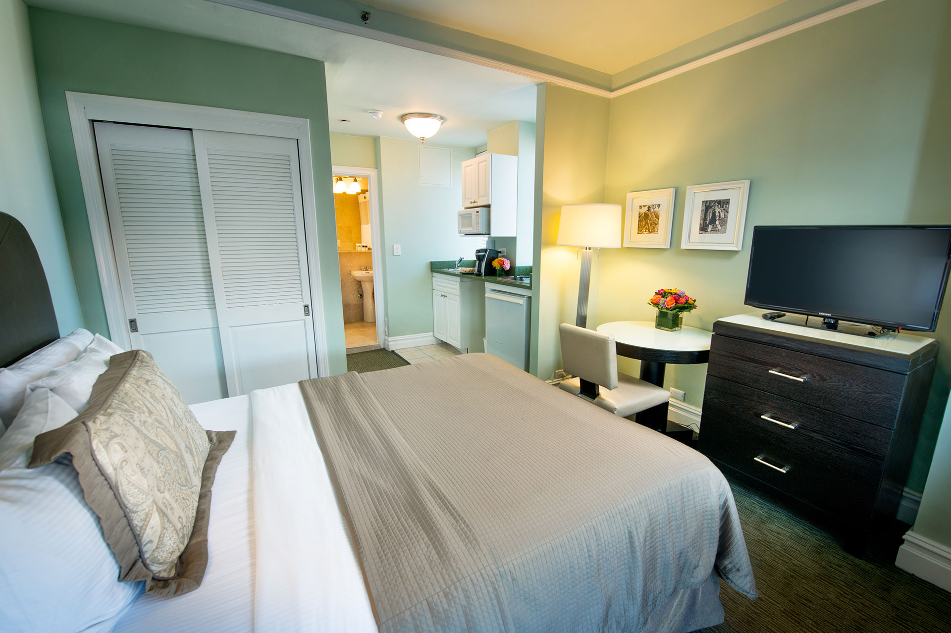 A wide view of a hotel room with a bed, TV, kitchenette, desk, and lamp.
